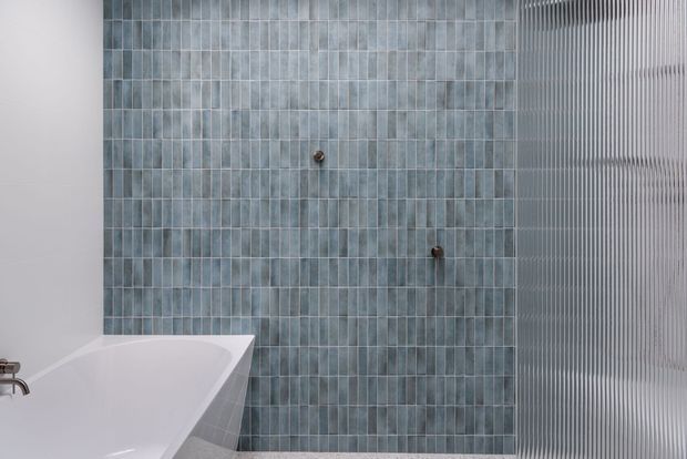 A minimalist bathroom featuring a white freestanding bathtub, a full-height wall of narrow blue-grey rectangular tiles, and a vertically ribbed translucent glass screen on the right. Brushed metal bath fittings are visible at the lower left, creating a cool, refined material palette.