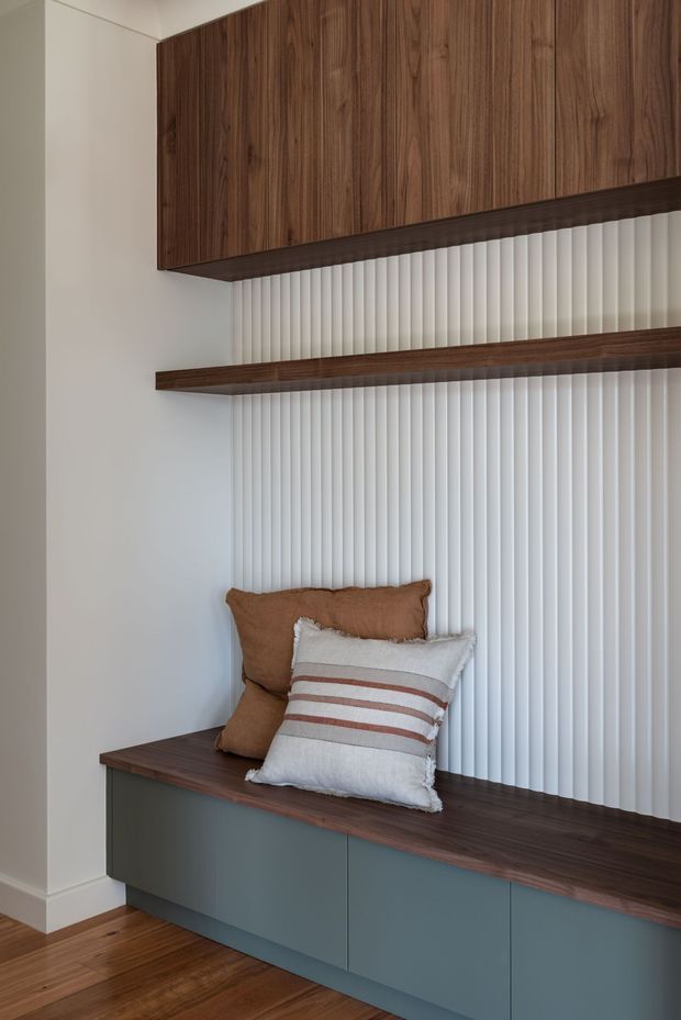 A built-in window seat with blue-grey cabinetry and a dark timber bench, framed by vertical white fluted wall panelling and matching dark timber overhead cabinetry and shelf. Neutral cushions add a warm, understated accent, while timber flooring continues the natural material palette.