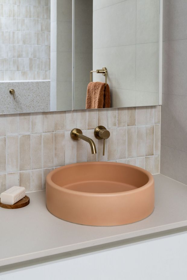 Close-up of a refined bathroom vanity featuring a large round terracotta vessel basin on a pale benchtop, wall-mounted brushed brass tapware, pale textured and rectangular ceramic tile splashback, mirrored shaving cabinet, and warm neutral styling.