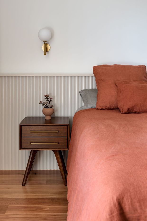 A warmly styled bedroom vignette featuring a mid-century-inspired dark timber bedside table with tapered legs and brass drawer pulls, positioned beside a bed dressed in terracotta bedding. The wall has vertical fluted panelling below a plain light wall, with a spherical wall sconce in a brass finish. Timber flooring and a small ceramic planter complete the scene.