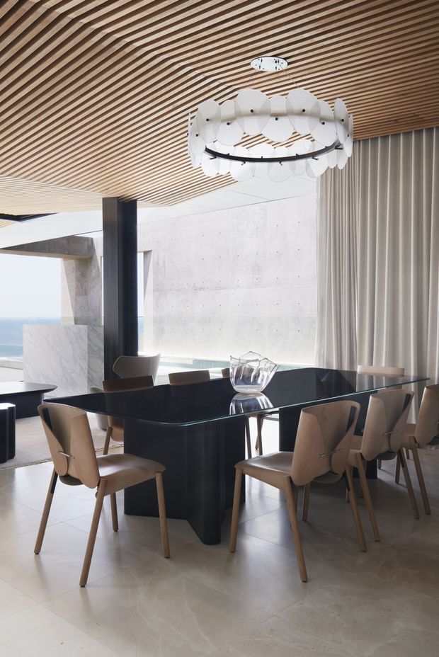 A refined contemporary dining area with a large black dining table surrounded by upholstered timber dining chairs. The room features a dramatic circular pendant light, a slatted timber ceiling, pale stone or concrete walls, floor-to-ceiling curtains, and expansive glazing with a coastal view beyond.