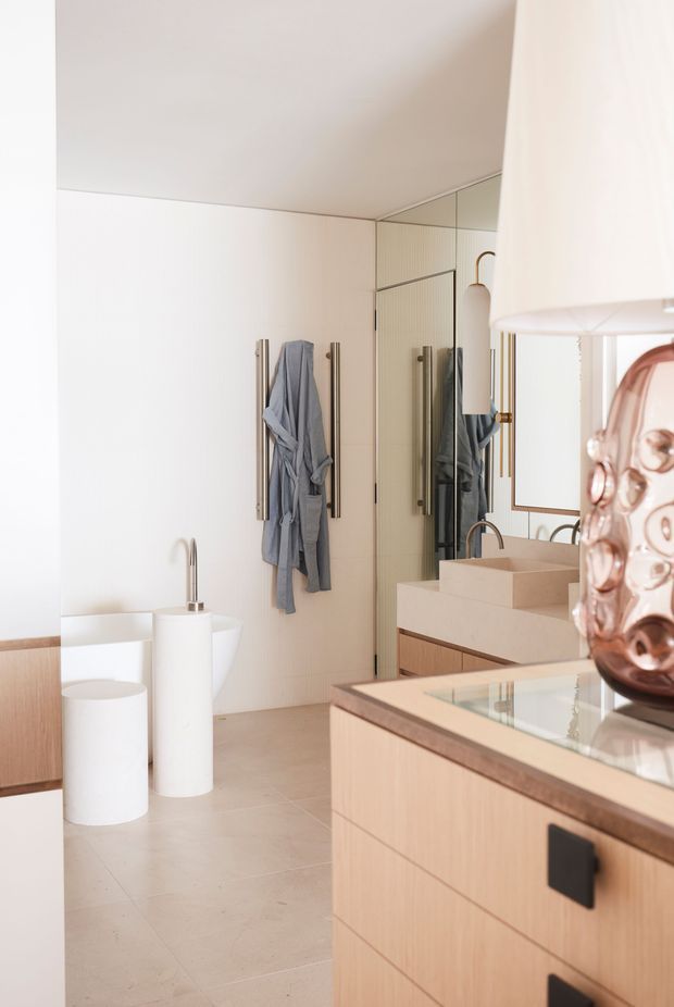 A bright, refined bathroom interior featuring a wall-mounted vanity with pale timber cabinetry, a thick beige stone benchtop and integrated basin, large mirrored cabinetry, and a freestanding white bathtub partially visible at left. Vertical towel rails hold a grey robe, while sculptural white and rose-toned decorative objects add a soft accent. The space has light tiled flooring, white walls, and a clean contemporary fit-out.