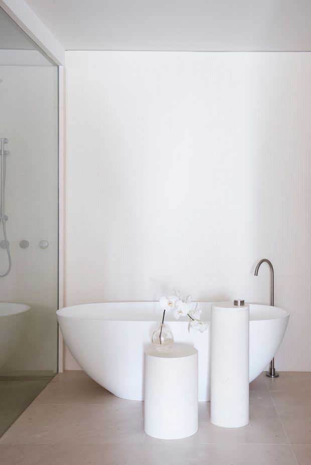 Minimalist contemporary bathroom featuring a freestanding white bathtub, walk-in glass shower, curved black tapware, cylindrical pedestal basins or display columns, and a white vertical-ribbed feature wall. The space has pale large-format tiled flooring, restrained white and beige tones, and a bright, serene atmosphere.