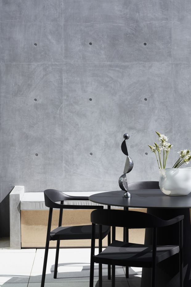 A refined dining area with a round black dining table and black chairs set against a pale grey concrete wall. A built-in upholstered bench runs along the wall, accompanied by a sculptural black-and-silver centerpiece and a white vase of flowers. Strong natural light creates crisp shadows across the light floor, producing a modern, minimalist atmosphere.