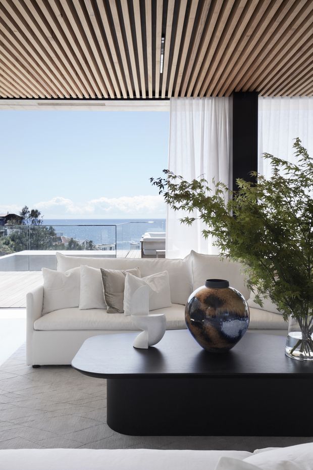 Bright, refined living room with a large white sectional sofa, sculptural coffee table, and dark rounded console or table. The room opens to a glass-balustraded terrace with ocean views, while sheer curtains, a black structural column, and a dramatic timber slatted ceiling add architectural interest. A large ceramic vessel and indoor tree provide natural accents.