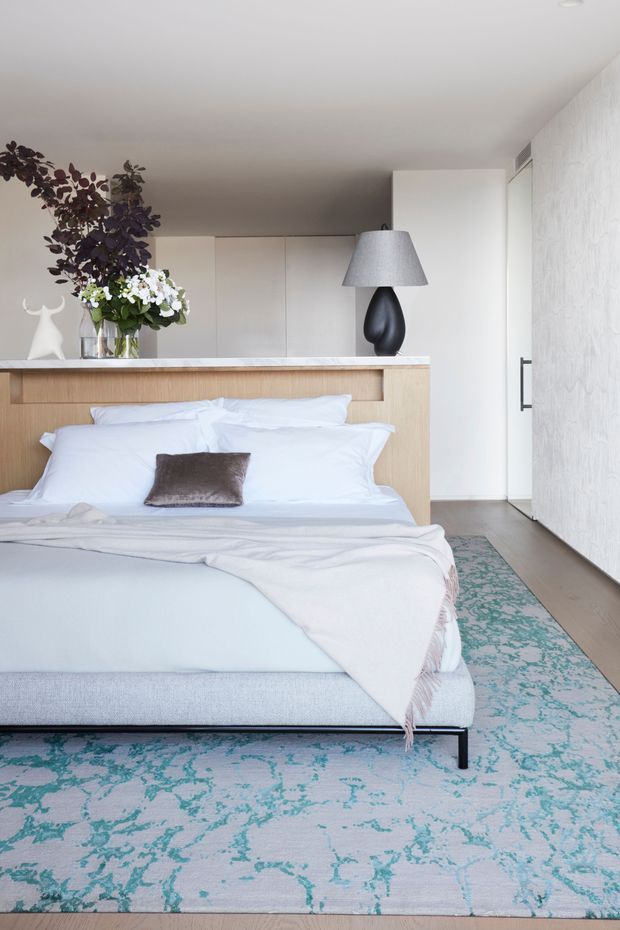 A bright, refined bedroom featuring a low upholstered bed dressed in white linens, a timber bedhead with integrated ledge, and a dark sculptural table lamp. Pale built-in cabinetry, light timber flooring, textured white wall treatment, and a large pale blue rug with turquoise pattern create a soft contemporary interior. Decorative flowers and a small sculptural object sit on the bedhead ledge.
