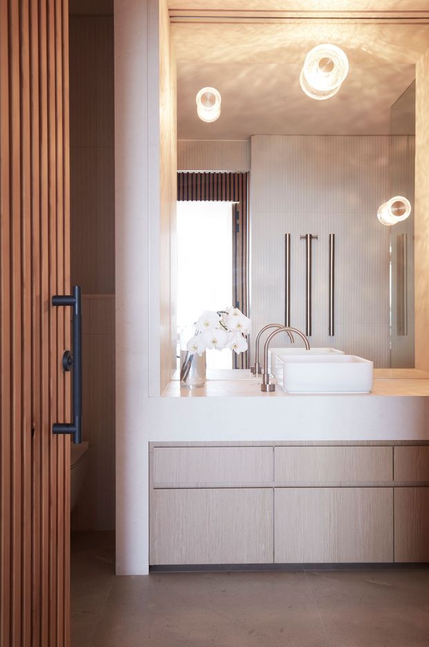 A refined contemporary bathroom vanity with a white vessel basin, brushed metal tapware, pale timber cabinetry, and a large mirrored wall. Vertical timber slats frame the left side, while ribbed wall panelling and integrated circular lighting add texture and interest. The warm timber and neutral palette create a calm, luxurious atmosphere.