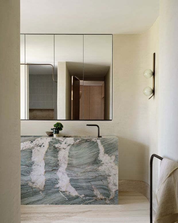 A refined minimalist bathroom with a floating or freestanding stone vanity clad in dramatic veined green-grey and white natural stone. A black tap, large wall mirror, globe wall lights, pale textured walls, and light stone flooring create a calm, sculptural interior. A towel rail is visible at right, with a glimpse of tiled shower or wet area reflected in the mirror.