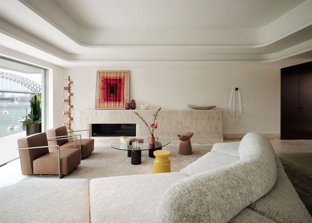 Bright, refined living room with a sweeping curved boucle sofa in the foreground, sculptural lounge chairs, layered round coffee tables, and a textured pale stone or tile fireplace surround. The room has a dramatic curved stepped ceiling, dark built-in cabinetry, large waterfront-facing glazing, and artful contemporary furnishings. A bridge structure and water are visible outside, while the overall interior is warm, neutral, and sophisticated.