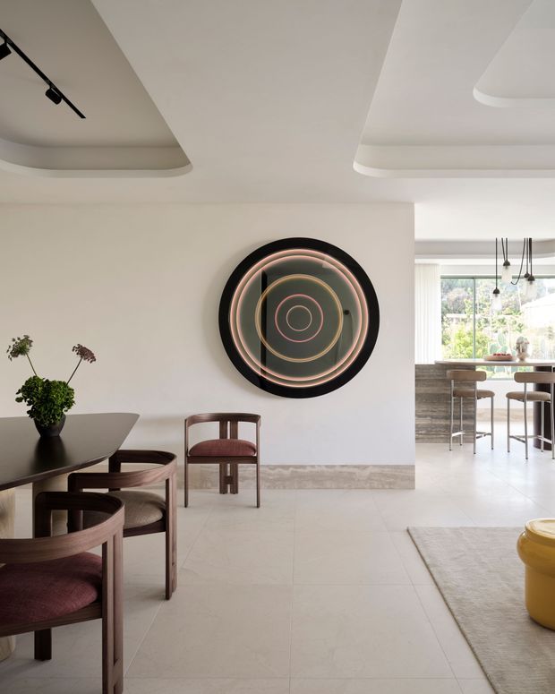 A refined open-plan dining and living area with a dark oval dining table and curved wooden chairs upholstered in muted burgundy fabric. The room has pale tiled flooring, warm off-white walls, sculptural recessed ceiling details, and a large circular abstract wall artwork. Through the opening, a bright kitchen or dining zone is visible with pendant lighting and extensive glazing overlooking greenery. A neutral rug and yellow accent stool appear in the living area.