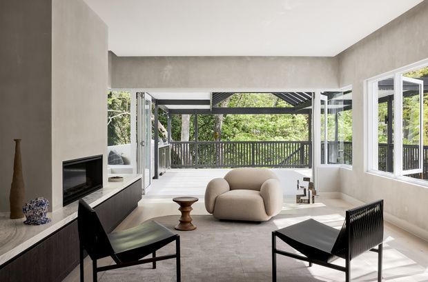 A bright contemporary living room with a minimalist concrete-look fireplace wall and built-in dark cabinetry. Two black lounge chairs and a sculptural cream armchair are arranged on a pale stone or concrete-look floor, opening onto a covered outdoor deck through extensive folding or sliding glazing. The deck has dark railings and a leafy outlook.