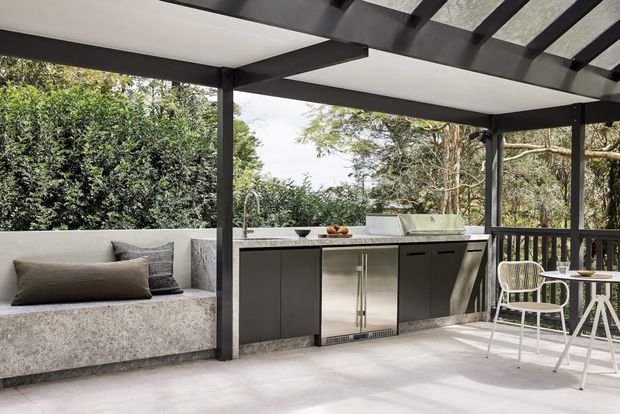 A covered outdoor entertaining area with a built-in outdoor kitchen, dark cabinetry, stainless-steel appliances and benchtop, and a concrete-look built-in bench seat. A small white dining table and chairs sit beside the kitchen, with lush greenery visible beyond the open sides. The space has a dark framed pergola roof with translucent panels and a relaxed contemporary feel.