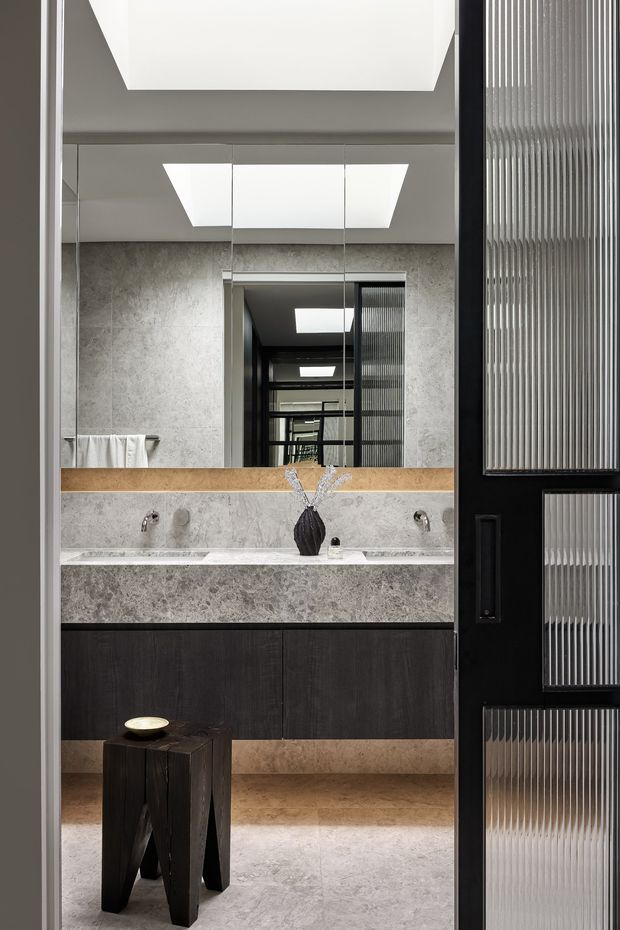 A refined contemporary bathroom viewed through a partially open black-framed door with ribbed translucent glass. The room features a wide double vanity with dark timber cabinetry, pale stone or terrazzo-like benchtop and matching wall and floor finishes, twin wall-mounted taps, a large mirror, and geometric ceiling skylights. A sculptural dark stool and small decorative object sit in the foreground.