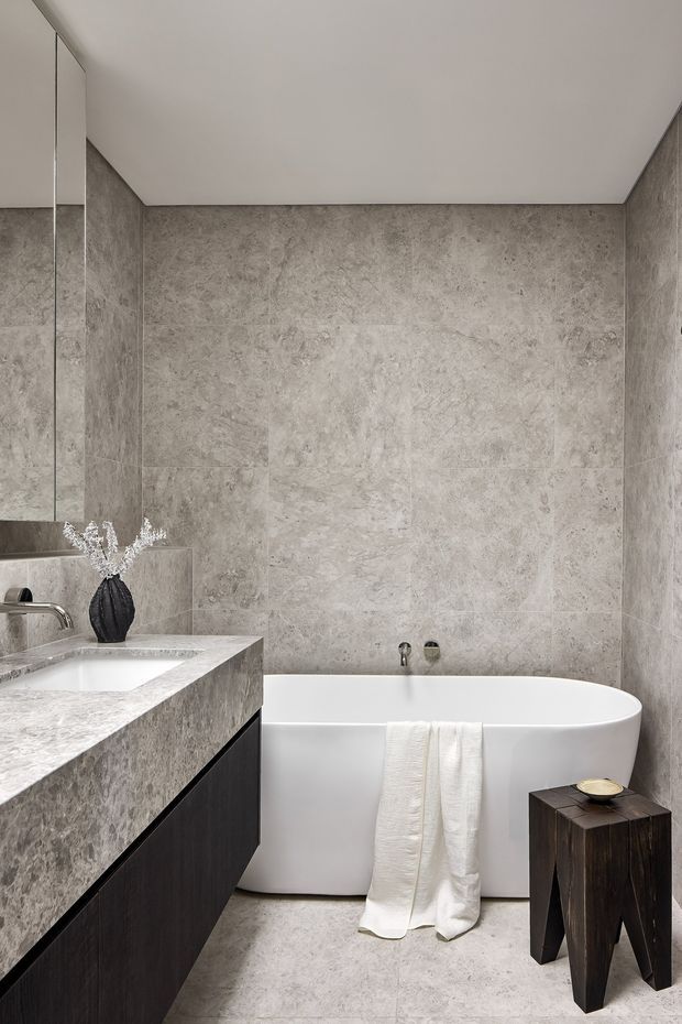 Contemporary bathroom with full-height grey stone-look wall and floor tiling, a freestanding white bathtub, wall-mounted dark timber vanity with a pale stone benchtop, and a large mirrored cabinet. The restrained neutral palette and clean lines create a refined, spa-like atmosphere.