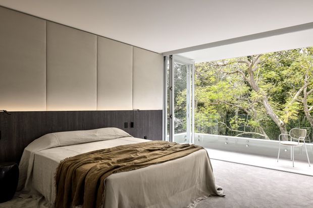 A contemporary bedroom with a low-profile bed dressed in neutral beige bedding and a brown throw. Floor-to-ceiling glazing opens onto a light-toned balcony or terrace overlooking mature trees. The room features full-height upholstered or padded wall panels above dark timber wall panelling, a pale carpeted floor, and a minimal, uncluttered fit-out.