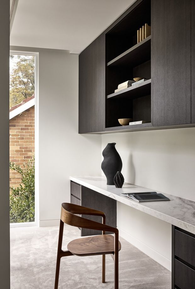 A refined built-in study nook or home office with dark timber cabinetry, open display shelving, a pale stone benchtop, and a sculptural wooden chair. Neutral plaster walls and carpet create a calm, minimalist setting, with a tall window bringing in natural light and views of a brick exterior.