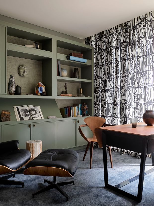 A sophisticated home office with built-in muted sage-green shelving and cabinetry, styled with books, ceramics, and artwork. A dark timber desk and mid-century leather lounge chair sit on a soft grey carpet, while full-height black-and-white patterned curtains add a bold graphic feature.