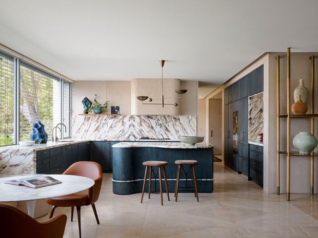 A refined open-plan kitchen and dining area with a dark island, dramatic veined marble splashback and benchtop, pale cabinetry, integrated dark timber storage, and a sculptural open shelving unit. Floor-to-ceiling windows with horizontal blinds bring in abundant natural light. A small round dining table and upholstered timber chairs sit in the foreground, while brass accents and contemporary pendant lights add warmth.