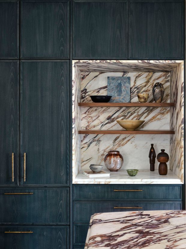 Close-up interior view of custom dark blue-black timber cabinetry with long brass-gold pulls, surrounding a marble-lined open display niche. The niche has dramatic white stone with burgundy and grey veining, timber shelves, and carefully arranged decorative ceramics and vessels. A matching veined stone countertop or island edge appears in the foreground.