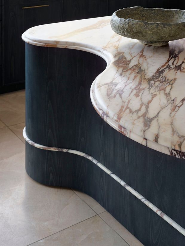 Close-up detail of a sculptural curved vanity or built-in counter with dark fluted timber cabinetry and a thick, organically shaped marble benchtop featuring prominent cream, taupe and burgundy veining. A rough stone vessel sits on the counter, and large pale floor tiles are visible below.