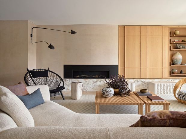 Warm, refined contemporary living room with a large textured cream sectional in the foreground, black woven lounge chair, sculptural black wall light, and a linear fireplace set into a pale stone or marble surround. Full-height light timber cabinetry spans the right wall, with open shelving displaying objects. A slatted timber coffee table, neutral patterned rug, soft beige plaster walls, and restrained natural decor create a calm, tactile interior.