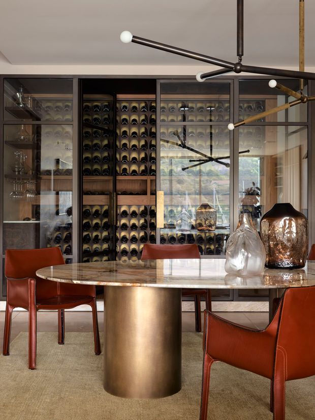 A refined dining room featuring a round marble-topped dining table with a cylindrical bronze pedestal, rust-red leather dining chairs, and a dramatic branching pendant light. The back wall is fitted with dark timber and glass wine storage cabinetry, with additional glassware display cabinets. Decorative blown-glass vessels sit on the table, and a large window reflection adds depth to the sophisticated, moody interior.