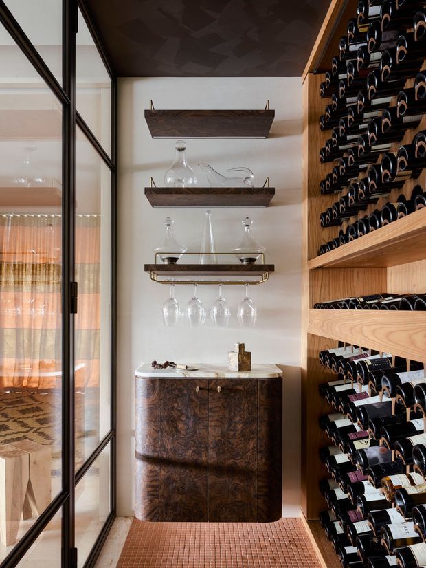 A refined compact wine storage and serving area with floor-to-ceiling timber wine racks densely filled with bottles on the right. A dark figured-wood drinks cabinet with a pale stone benchtop sits beneath floating timber shelves displaying decanters and glassware, while a framed glass partition reveals an adjacent room. The space has warm, luxurious detailing and a patterned ceiling.