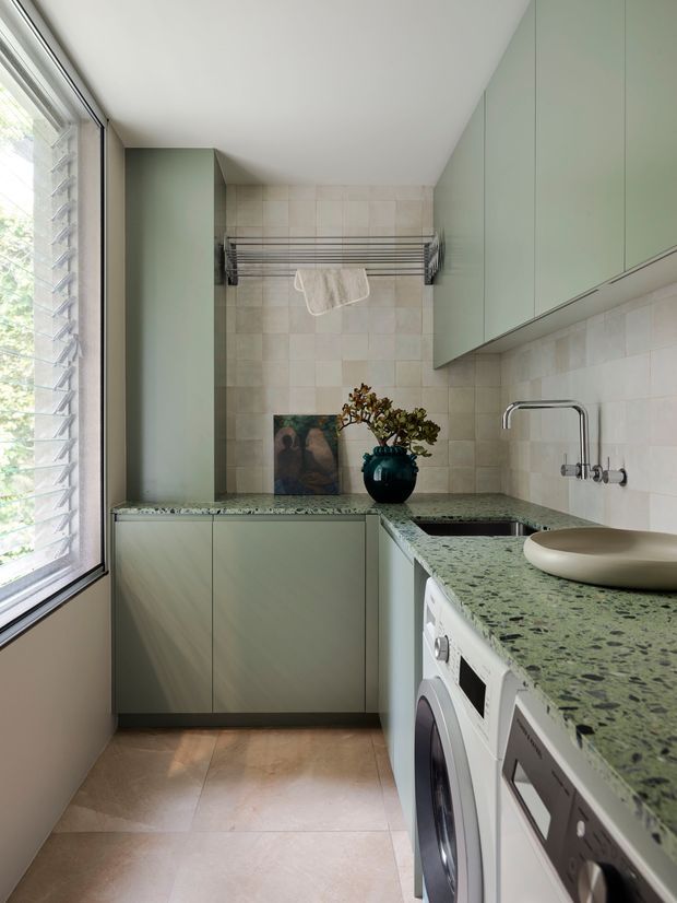 A compact, bright laundry room with pale sage-green full-height cabinetry, a green-speckled stone benchtop, pale tiled walls and floor, integrated washing appliances, a stainless-steel sink and tap, and a wall-mounted drying rack. Louvered glazing along the left side provides natural light. The room has a clean, refined contemporary appearance with soft natural colours.