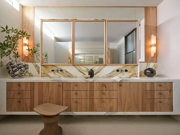 A refined double-vanity bathroom with a wide floating timber vanity, extensive drawers, a pale stone benchtop and dramatic veined stone splashback. Three framed mirrors sit between timber wall panels, with sculptural wall sconces on either side. Decorative vessels and a small timber stool add warmth to the calm, contemporary space.