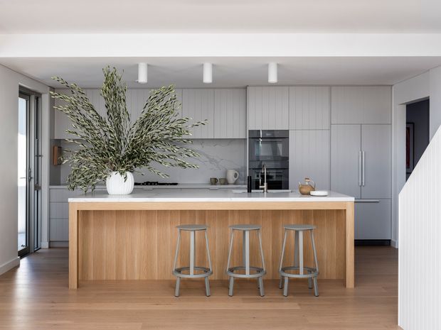 A bright, refined contemporary kitchen featuring a large island with a light timber panelled base and white benchtop, three metal bar stools, and full-height pale grey cabinetry. Integrated black appliances, a slim linear rangehood, white cylindrical pendant-style ceiling lights, and warm timber flooring complete the clean, open-plan space. A large leafy arrangement provides a natural focal point.