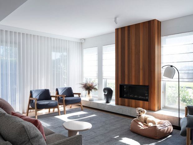 Bright contemporary living room with a large grey sectional sofa, two timber-framed lounge chairs, a round white coffee table, and a dog bed. Floor-to-ceiling sheer window coverings and multiple windows provide abundant natural light. A dramatic vertical timber slat feature wall incorporates a black built-in fireplace or media unit, with a long low stone-like hearth below.