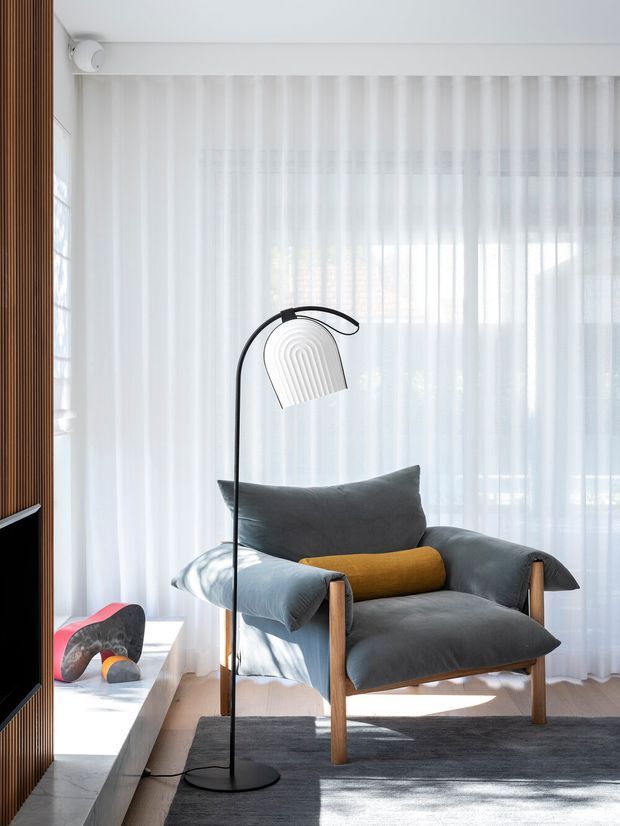 Bright, refined sitting area featuring a grey upholstered lounge chair with wooden arms, a mustard cylindrical cushion, and a sculptural black floor lamp. Sheer white curtains span the window wall, while a vertical timber slat feature and low white display ledge frame the space. A dark rug and pale timber floor complete the contemporary interior.