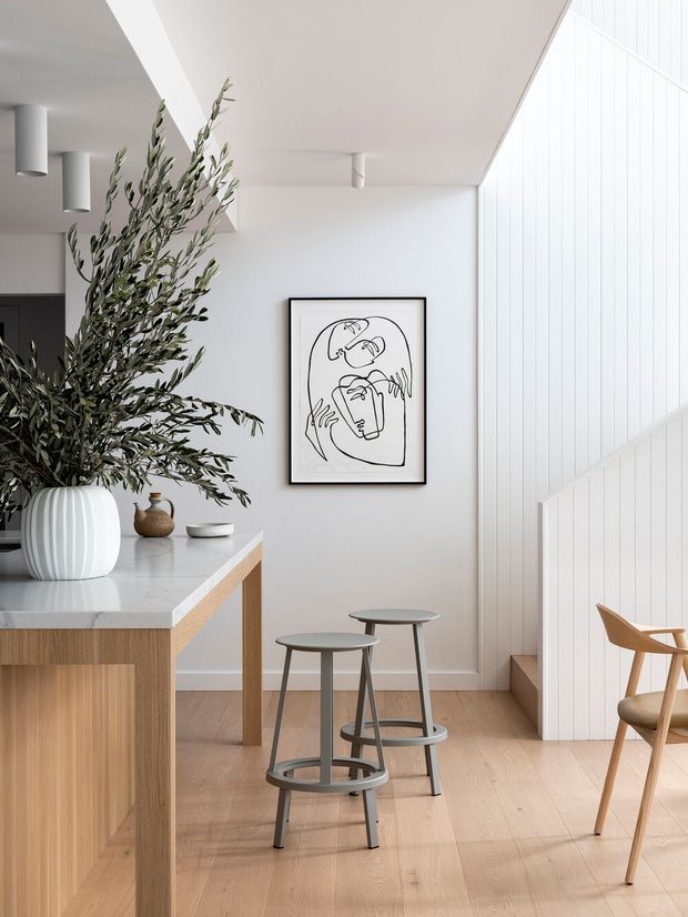 Bright contemporary kitchen and dining area with a timber island, pale stone benchtop, light timber flooring, minimalist stools, and a sculptural timber chair. White vertical wall panelling and a stair enclosure create a clean architectural backdrop, complemented by a large olive branch arrangement and monochrome line artwork.