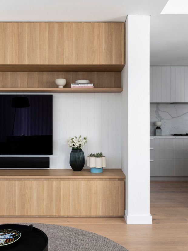 A refined open-plan living room featuring a built-in timber entertainment unit with upper cupboards, open shelving and a low media cabinet. White vertical wall panelling provides a backdrop for the television and decorative objects, while a black coffee table and textured rug sit in the foreground. A contemporary kitchen with grey cabinetry and a marble-look splashback is visible beyond the dividing wall.