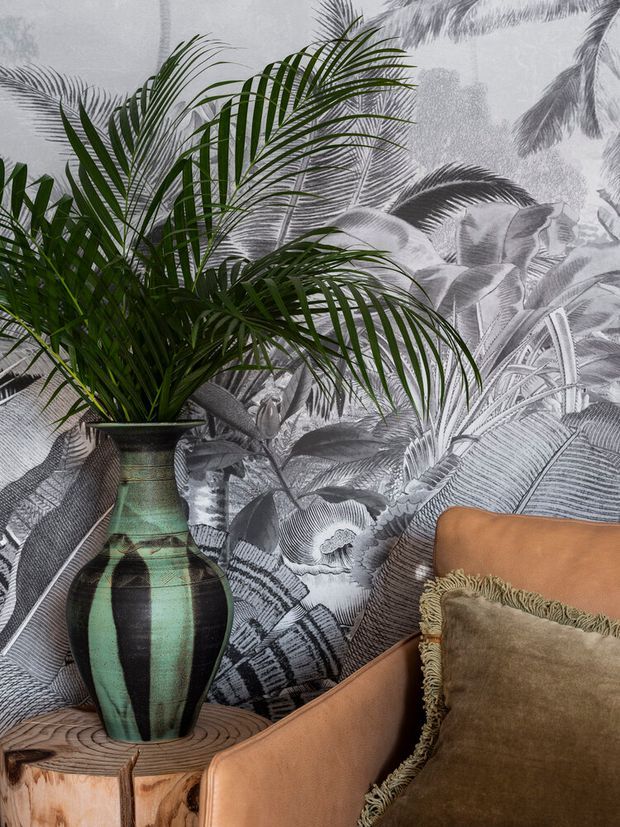 A close-up interior vignette featuring a large green and black ceramic vase with a palm plant, placed on a rustic timber side table beside a tan leather armchair. The wall behind is covered in dramatic monochrome tropical botanical wallpaper, complemented by a textured olive-brown fringed cushion. The scene has an eclectic, richly layered tropical aesthetic.
