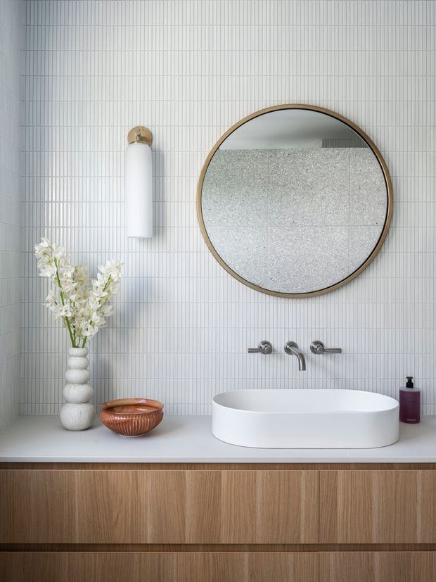 A refined contemporary bathroom vanity with a freestanding white vessel basin, wall-mounted brushed metal tapware, a large round timber-framed mirror, and a ribbed pale wall tile backdrop. The vanity features a light timber-grain floating cabinet with a pale solid countertop. Decorative flowers, a ceramic bowl, wall sconce, and soap dispenser add restrained styling.