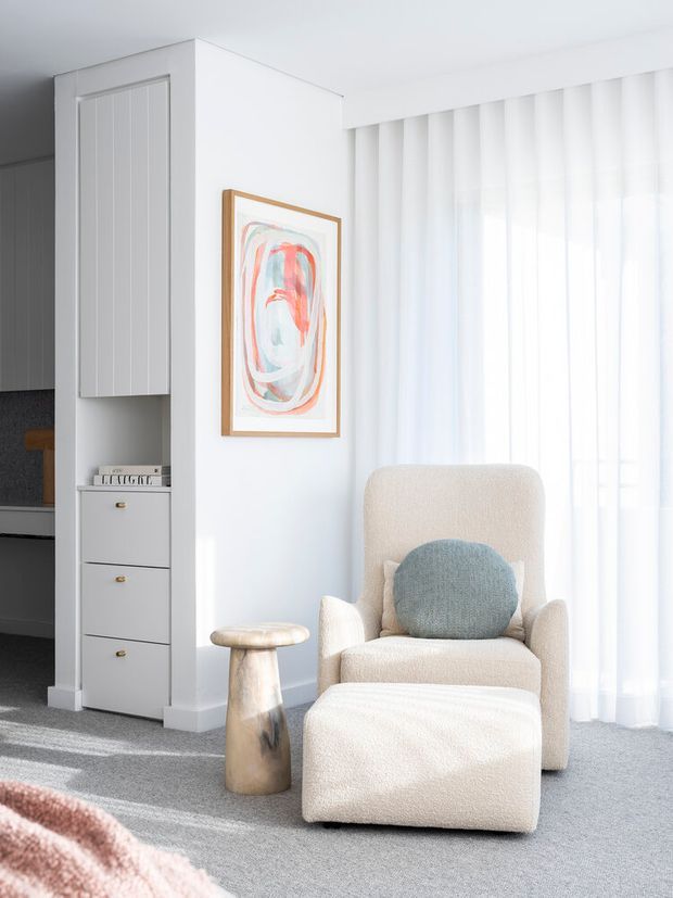 Bright, softly styled seating nook with a cream boucle armchair and matching ottoman, a pale blue accent cushion, a small sculptural side table, abstract artwork, sheer floor-to-ceiling curtains, and integrated white cabinetry with drawers. The space has a clean, serene, contemporary feel with abundant natural light.