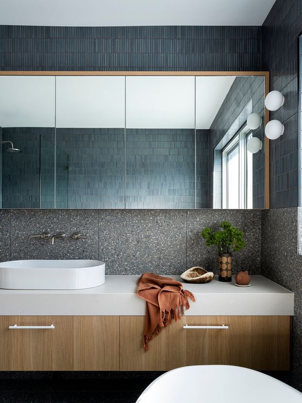 A sophisticated contemporary bathroom featuring a wall-mounted timber vanity with a white solid-surface benchtop, a white vessel basin, and dark vertically textured wall tiles. A large timber-framed mirror spans the vanity, reflecting a glass shower enclosure and tall window. Spherical wall lights, dark flooring, and restrained natural accessories complete the refined scheme.