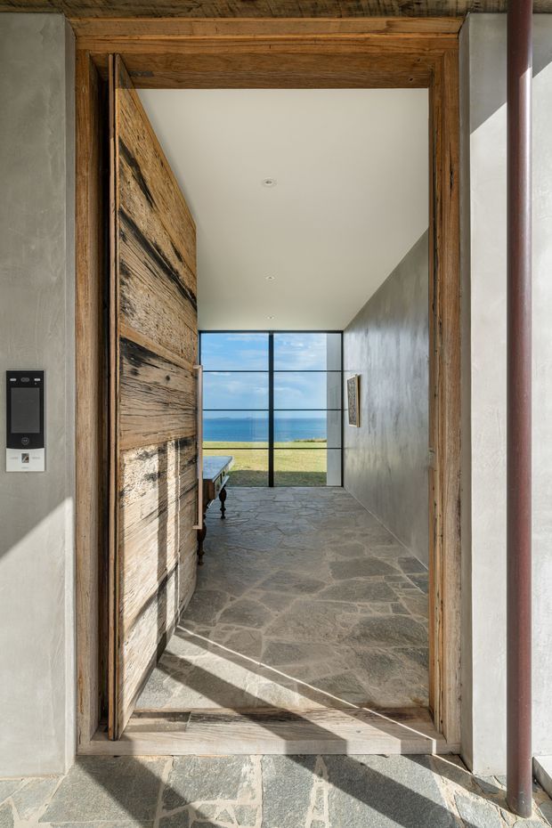 A sunlit entryway viewed through a substantial reclaimed timber doorway, opening into a long circulation space with a rough natural-stone floor, plastered walls, and a full-height black-framed glazed window at the far end. The glazing looks toward a grassy coastal landscape and ocean horizon. The scene combines rustic tactile materials with clean contemporary detailing.