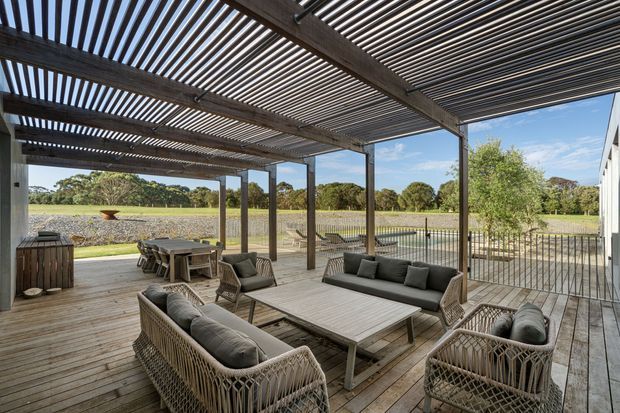 A spacious covered outdoor entertaining area with a timber deck and slatted pergola roof. Wicker lounge seating, sofas, armchairs, a low coffee table, and an outdoor dining table are arranged beneath the pergola, overlooking a landscaped rural or park-like setting. The space has a relaxed contemporary coastal feel, with dark timber framing, open sides, and a metal balustrade.