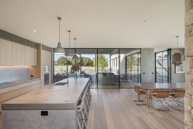 A spacious open-plan contemporary kitchen and dining area with a long pale stone island, light timber cabinetry, integrated appliances, pendant lights, and timber flooring. Full-height black-framed glazing connects the interior to an outdoor area, while a substantial exposed stone wall forms the right edge of the room. A dining table and woven chairs sit beside the kitchen.