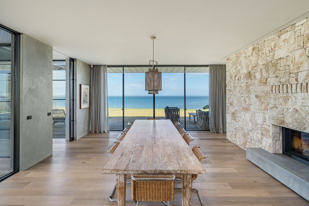 A spacious contemporary dining room with a long rustic timber dining table and woven dining chairs, opening through full-height glazing to a beachfront or ocean view. The room features pale timber flooring, a large natural stone feature wall with an integrated fireplace, sheer grey curtains, dark-framed glazing, and a woven pendant light. An outdoor terrace and ocean are visible beyond the dining area.