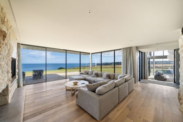 A spacious contemporary living room with a large neutral sectional sofa and round coffee table, opening through floor-to-ceiling corner glazing onto a timber deck and expansive ocean and coastal landscape views. The room features wide-plank timber flooring, pale walls and ceiling, full-height curtains, and prominent rough natural-stone wall elements at both sides.
