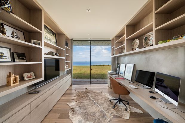 A contemporary home office lined with extensive built-in timber shelving and cabinetry. A long desk with multiple computer monitors runs along the right wall, while display shelving, framed artwork, books and decorative objects occupy both sides. A large sliding glass door at the far end opens toward a grassy coastal outlook. The room has light timber flooring, a pale textured wall behind the desk, recessed ceiling lighting, and a hide rug, creating a refined, warm workspace.