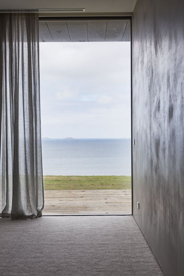 A minimalist interior looking through a full-height glazed sliding door toward a timber deck, grassy shoreline and calm ocean horizon. The room has grey carpet, a dark textured concrete-like wall, sheer grey curtains, and a slim linear ceiling vent or slot. The composition emphasises the strong connection between the subdued interior and the coastal outlook.