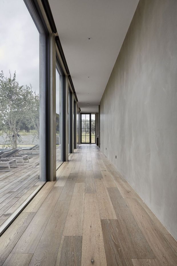 A long, minimalist interior hallway with wide pale timber floorboards, a smooth light plaster wall, and full-height dark-framed glazing along one side. The glazing opens views to an outdoor timber deck and landscaped surroundings, while recessed ceiling lights and a glazed door at the far end emphasize the corridor's linear perspective.