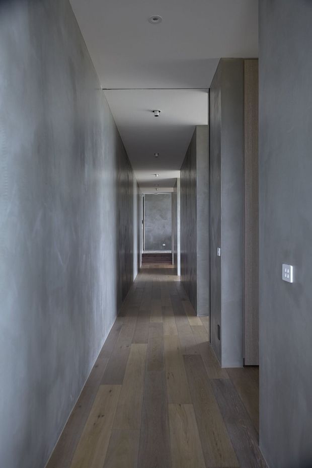 A long, narrow modern hallway with continuous grey textured walls, a white ceiling with recessed and small surface-mounted lights, and wide-plank timber flooring. Full-height dark grey and timber-toned doors or built-in panels line the right side, leading toward a brighter end of the corridor.