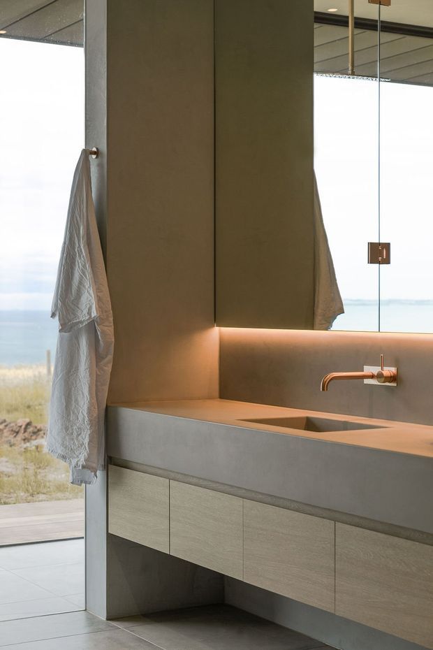 A refined minimalist bathroom vanity with a long integrated concrete or stone benchtop and inset basin, wall-mounted brushed copper tapware, warm under-cabinet lighting, and pale wood-grain floating cabinetry. A large mirror reflects the ceiling and pendant detail. Full-height glazing opens to a bright coastal landscape, while a white textured towel hangs beside the vanity.
