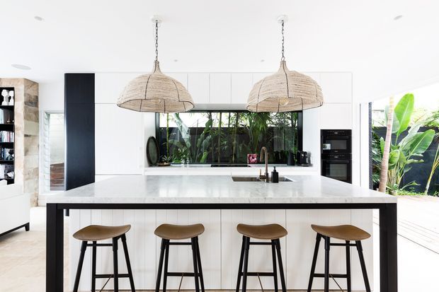 Bright contemporary kitchen centred on a large white island with seating for four. The space features white flat-panel cabinetry, a pale stone benchtop, black structural and appliance accents, timber bar stools, woven pendant lights, and a glazed opening with lush greenery beyond.