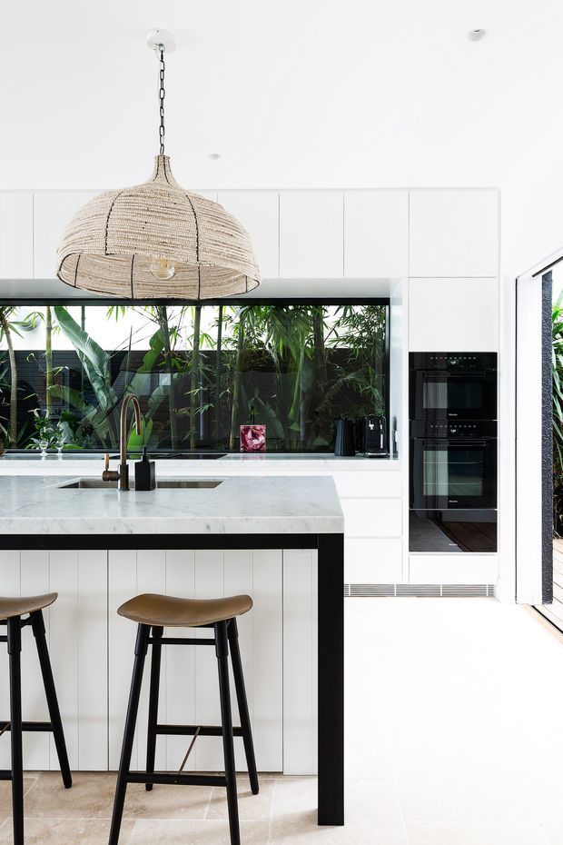 Bright contemporary kitchen featuring a large white marble island with integrated sink, white vertical-panel cabinetry, black-framed glazing overlooking lush greenery, and a tall bank of built-in black ovens. A woven pendant light hangs above the island, with two timber-and-black bar stools in the foreground.